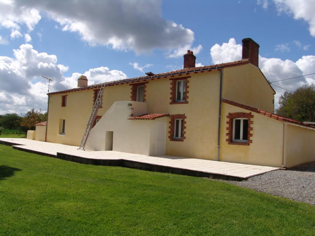 Maison jaune à briques rouges avec terrasse et échelle, jardin verdoyant.
