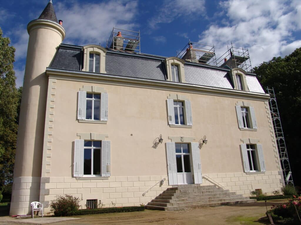 Manoir avec tourelle et volets blancs, échafaudages sur le toit en ardoise sous un ciel bleu.