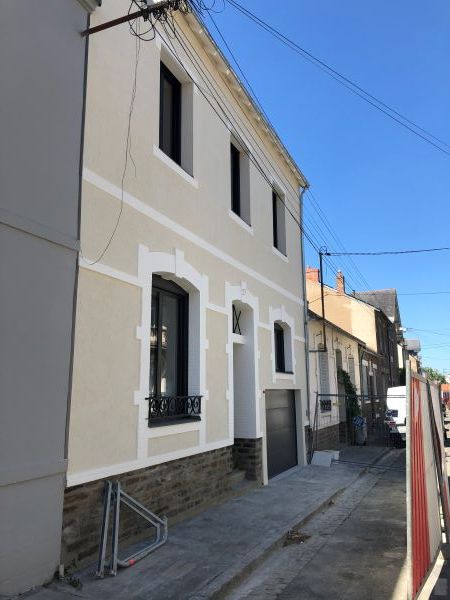Façade de maison beige rénovée, encadrements blancs, soubassement pierre et garage.