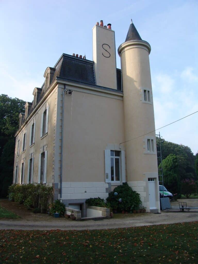 Manoir beige avec tourelle, toit à la Mansart et grande cheminée marquée d'un S sous un ciel bleu.