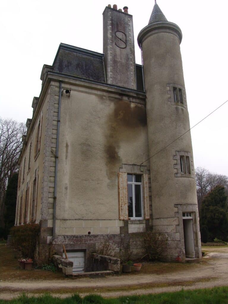 Manoir en pierre avec tour ronde et cheminée ornée d'un S sous un ciel couvert.