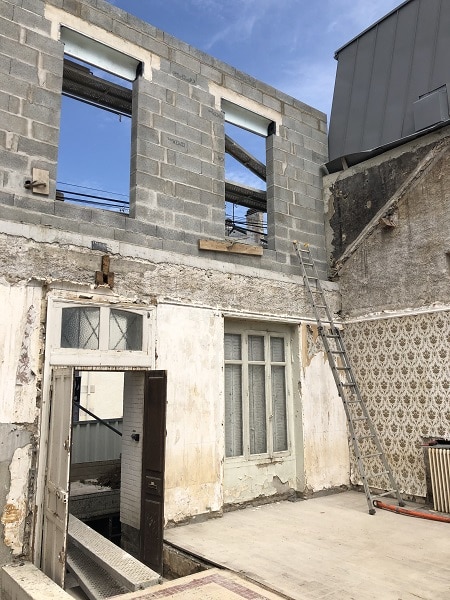 Rénovation de bâtiment, mur en parpaings sur structure ancienne, échelle et fenêtres ouvertes sous ciel bleu.